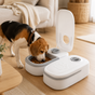 Dog eating from a white pet food dispenser in a home setting.