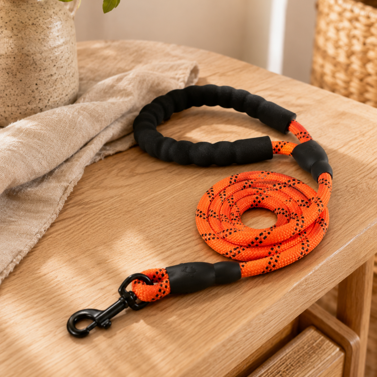 Black and orange braided dog leash on a wooden surface with a plant and woven basket in the background.